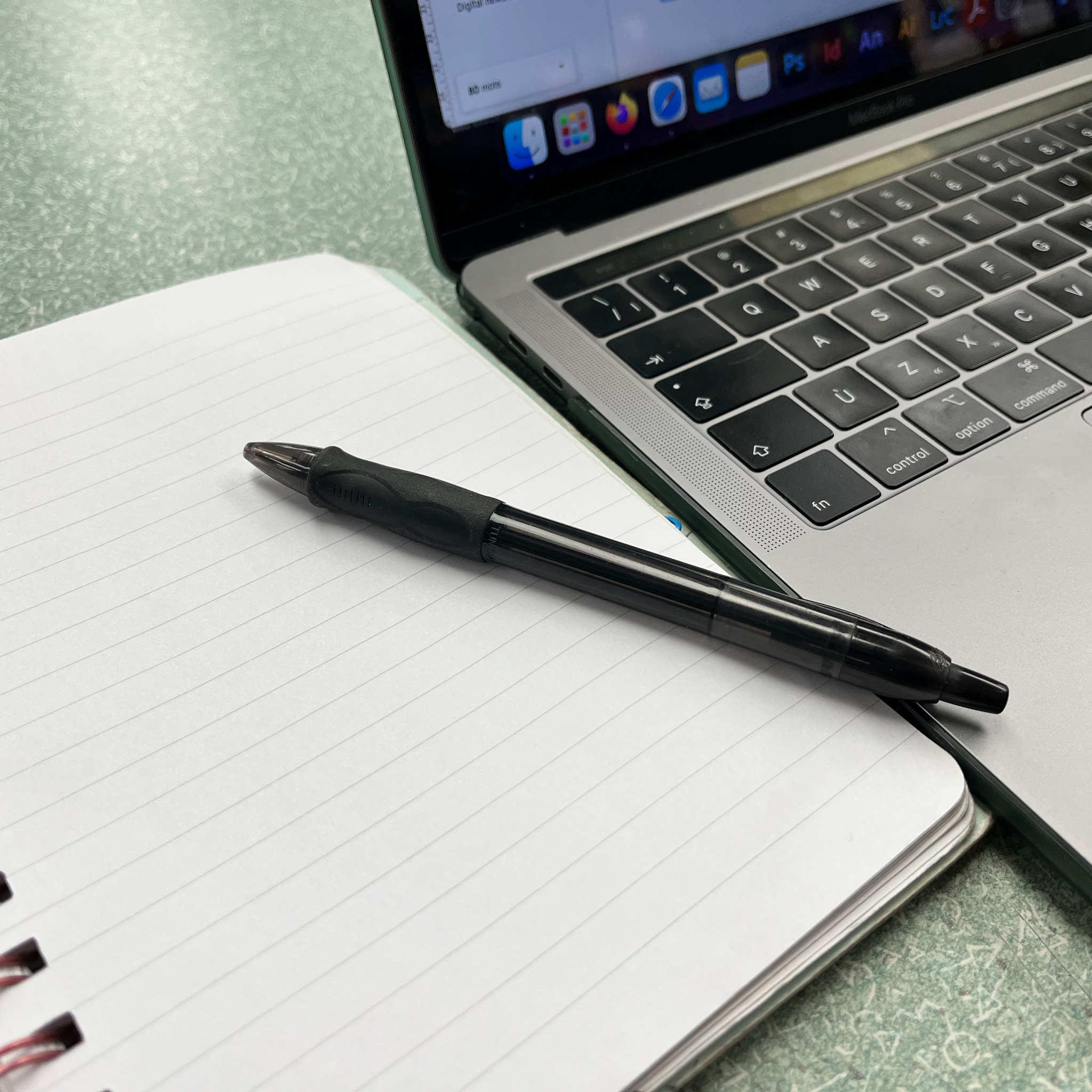 A cropped image of a computer and a notebook with a pen on top of it.